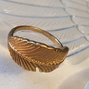 May include: A gold-toned ring with a feather design. The feather is detailed with lines and has a slightly textured surface.
