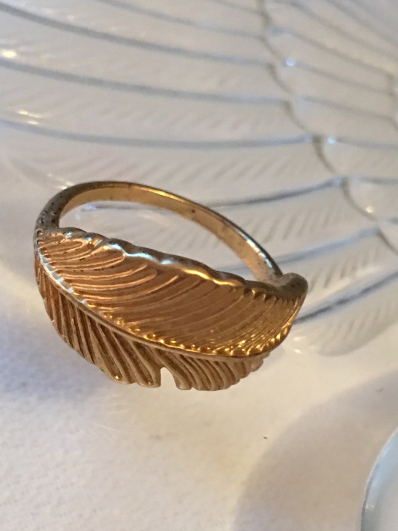 May include: A gold-toned ring with a feather design. The feather is detailed with lines and has a slightly textured surface.