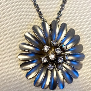 Könnte beinhalten: Halskette mit silberfarbenem Anhänger in Form einer Gänseblume. Der Anhänger hat eine große Blume mit mehreren Blütenblättern und kleinen, klaren Kristallen in der Mitte. Die Kette ist ebenfalls silberfarben.