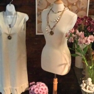 May include: Two white sleeveless dresses on display. One dress is on a mannequin and the other is hanging on a hanger. Both dresses have a ruffled hem. The mannequin is wearing multiple necklaces.