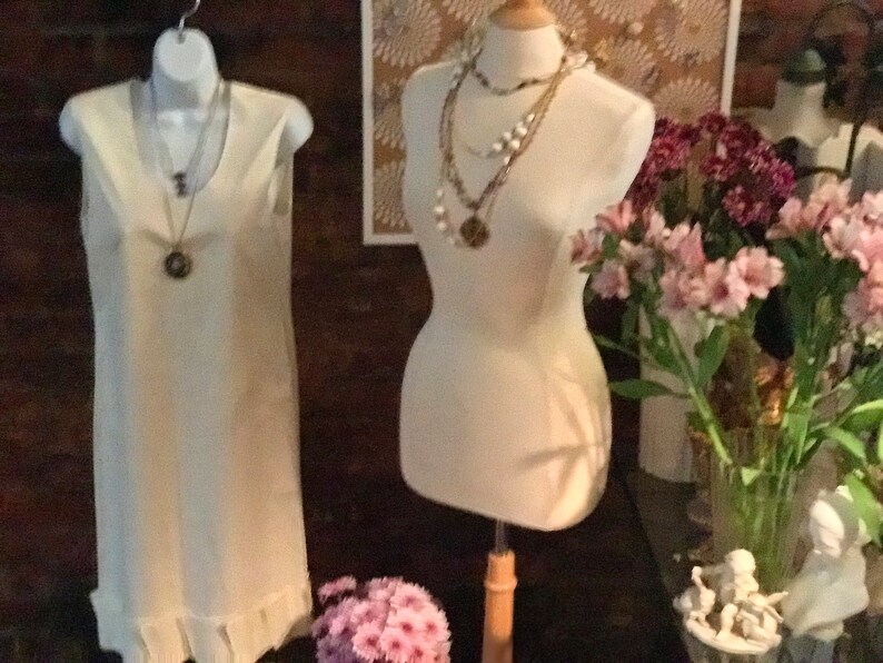 May include: Two white sleeveless dresses on display. One dress is on a mannequin and the other is hanging on a hanger. Both dresses have a ruffled hem. The mannequin is wearing multiple necklaces.