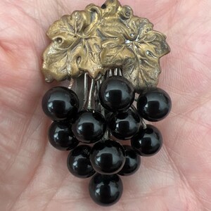 May include: A vintage brooch featuring a cluster of black glass grapes hanging from a gold-toned metal vine with leaves.