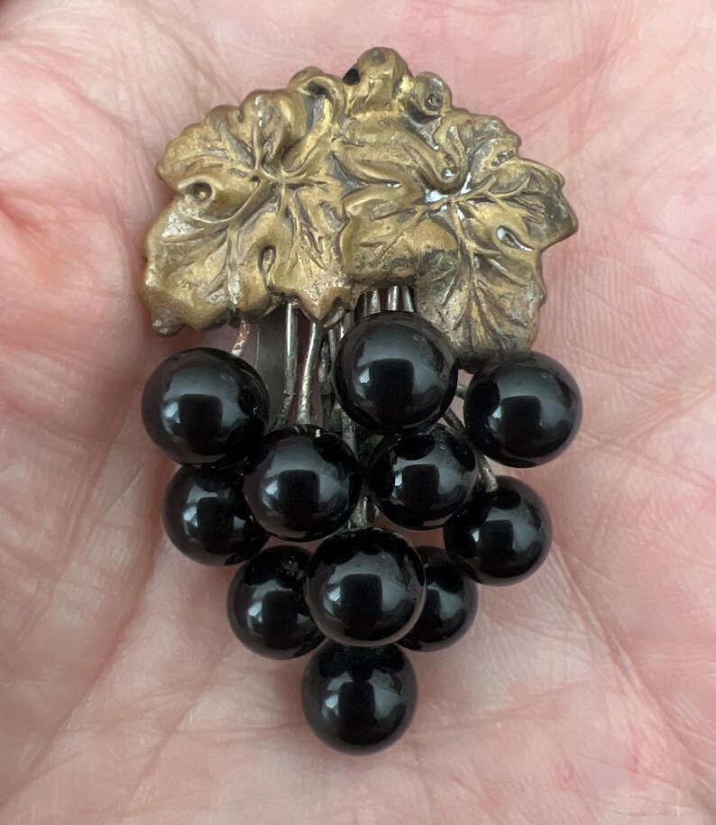 May include: A vintage brooch featuring a cluster of black glass grapes hanging from a gold-toned metal vine with leaves.