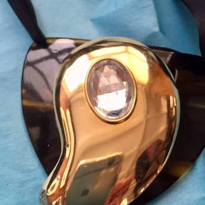 May include: A gold-toned pendant with a clear crystal in the center, hanging from a black ribbon.