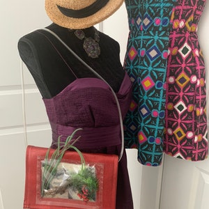 May include: A red crossbody bag with a clear plastic window showcasing a small potted plant. The bag has a strap and is worn on a mannequin with a purple dress and a straw hat.