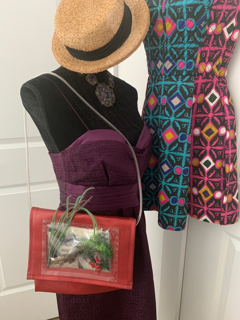 May include: A red crossbody bag with a clear plastic window showcasing a small potted plant. The bag has a strap and is worn on a mannequin with a purple dress and a straw hat.