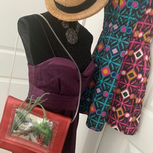 May include: A red purse with a clear plastic window containing a small potted plant. The purse has a long strap and is worn over the shoulder. The purse is displayed on a mannequin with a purple dress and a straw hat.