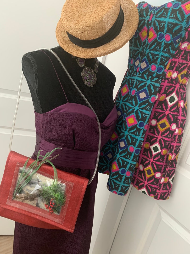 May include: A red purse with a clear plastic window containing a small potted plant. The purse has a long strap and is worn over the shoulder. The purse is displayed on a mannequin with a purple dress and a straw hat.