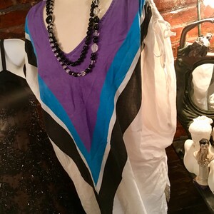 May include: A white, purple, blue and black striped tunic top with a v-neck and a beaded necklace.