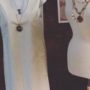 May include: A white sleeveless dress with a ruffled hem and a silver necklace with a large, round pendant. The dress is displayed on a white mannequin.