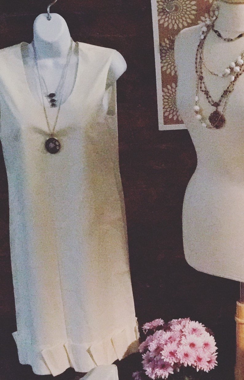 May include: A white sleeveless dress with a ruffled hem and a silver necklace with a large, round pendant. The dress is displayed on a white mannequin.