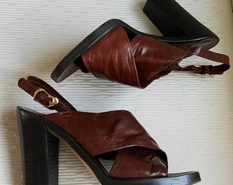 May include: A pair of brown leather heeled sandals with a criss-cross strap design. The sandals have a chunky black heel and an ankle strap with a gold buckle.