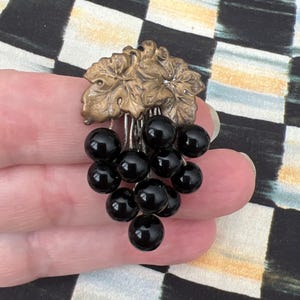 May include: A gold-toned grapevine cluster with black glass beads hanging from it. The cluster is on a hand.