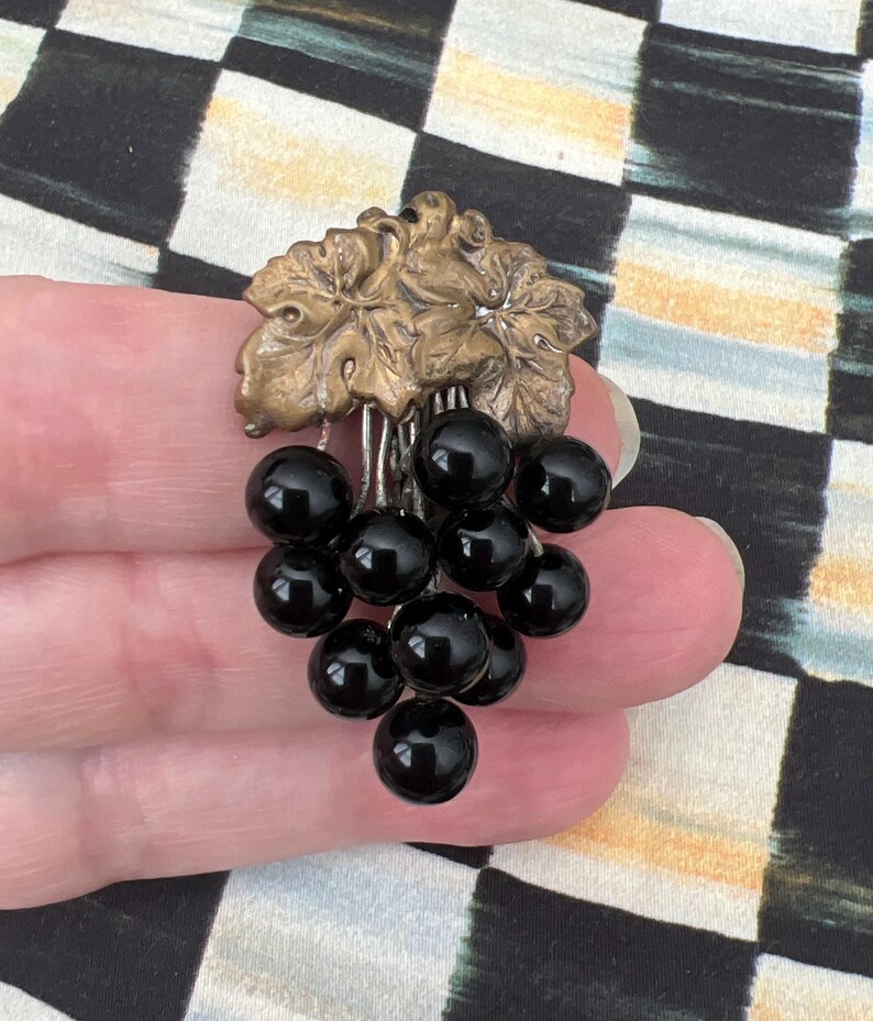 May include: A gold-toned grapevine cluster with black glass beads hanging from it. The cluster is on a hand.