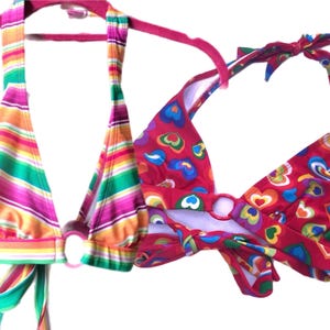 Op de afbeelding: Twee bikinitops met halterstijl. Eén is gestreept met groen, roze, oranje en geel. De andere is rood met een kleurrijk hartpatroon. Beide hebben ringdetails en striksluitingen.