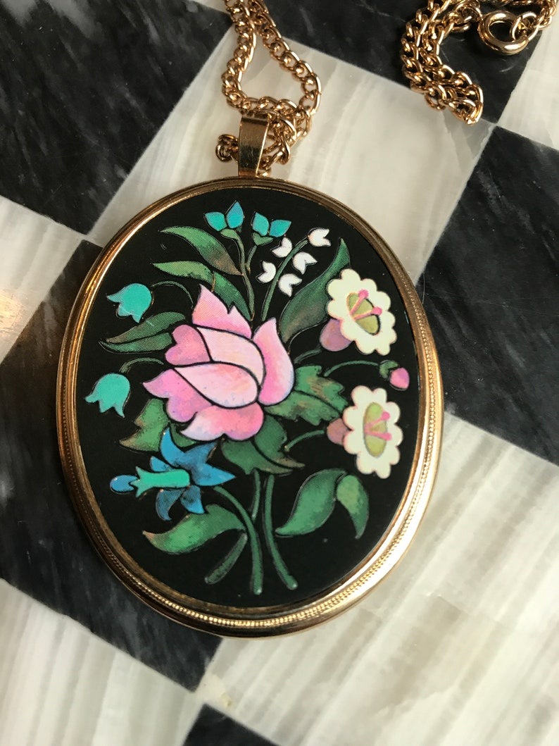 May include: A gold chain necklace with an oval pendant featuring a black background and a colorful floral design. The pendant has a pink rose, white flowers, blue flowers, and green leaves.