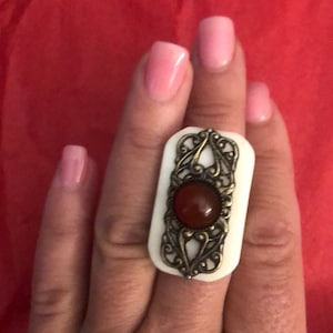 Könnte beinhalten: Ein weißer Ring mit einem goldenen Filigranmuster und einem roten Edelstein in der Mitte.