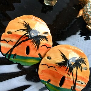 May include: Two painted sand dollar ornaments with a sunset scene and palm trees. The sand dollars are painted with orange, yellow, green, and blue colors.