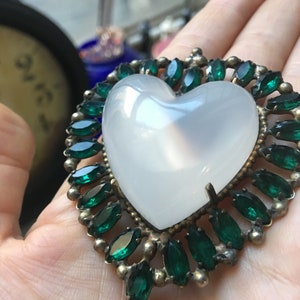 May include: A heart-shaped pendant with a large, opaque white center stone. The pendant is framed by a border of small, emerald-green gemstones set in a gold-toned metal. The design is vintage and elegant.