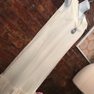 May include: A white sleeveless dress with a ruffled hem. The dress is on a mannequin and has a gold chain necklace with a blue and white cameo pendant.