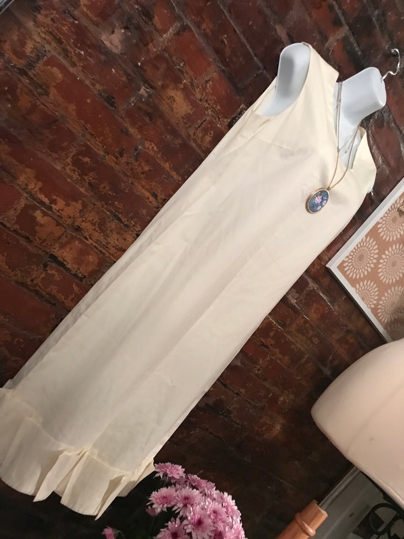 May include: A white sleeveless dress with a ruffled hem. The dress is on a mannequin and has a gold chain necklace with a blue and white cameo pendant.