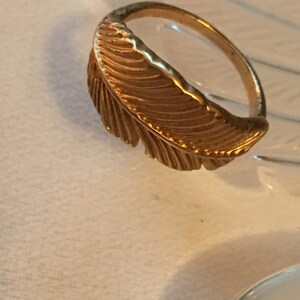 May include: A gold-toned ring with a feather design. The feather is detailed with lines and curves.