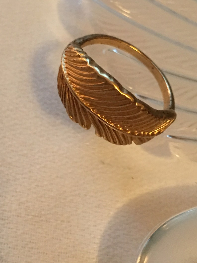 May include: A gold-toned ring with a feather design. The feather is detailed with lines and curves.