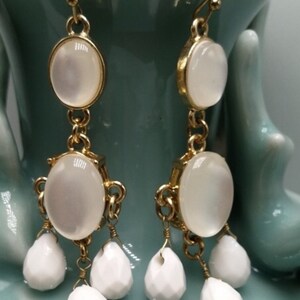 May include: Gold-tone dangle earrings with white, oval-shaped stones and teardrop-shaped accents. The earrings have a classic design with a delicate chain and hook closure. The earrings are set against a light blue background.