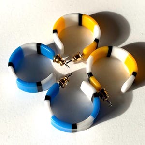May include: Four pairs of colorful hoop earrings. Each earring features a curved design with a combination of white, blue, yellow, and black stripes. The earrings have gold-colored posts.