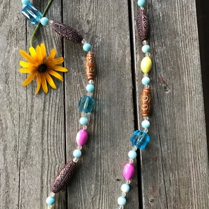 May include: A colorful beaded necklace with brown, pink, yellow, and blue beads. The necklace is made of various materials, including wood and glass. The necklace is on a wooden surface.