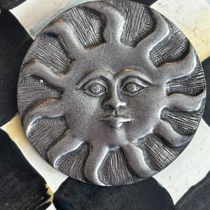 May include: A silver-toned metal sun with a face and rays. The sun is a decorative object with a detailed design.