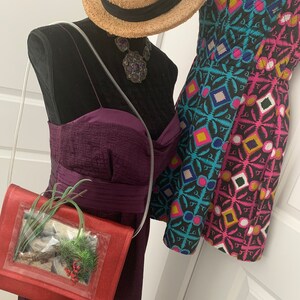 May include: A red purse with a clear plastic window showing a small potted plant inside. The purse is on a mannequin wearing a purple dress and a straw hat with a black band. The mannequin is also wearing a large, dark purple and silver necklace.