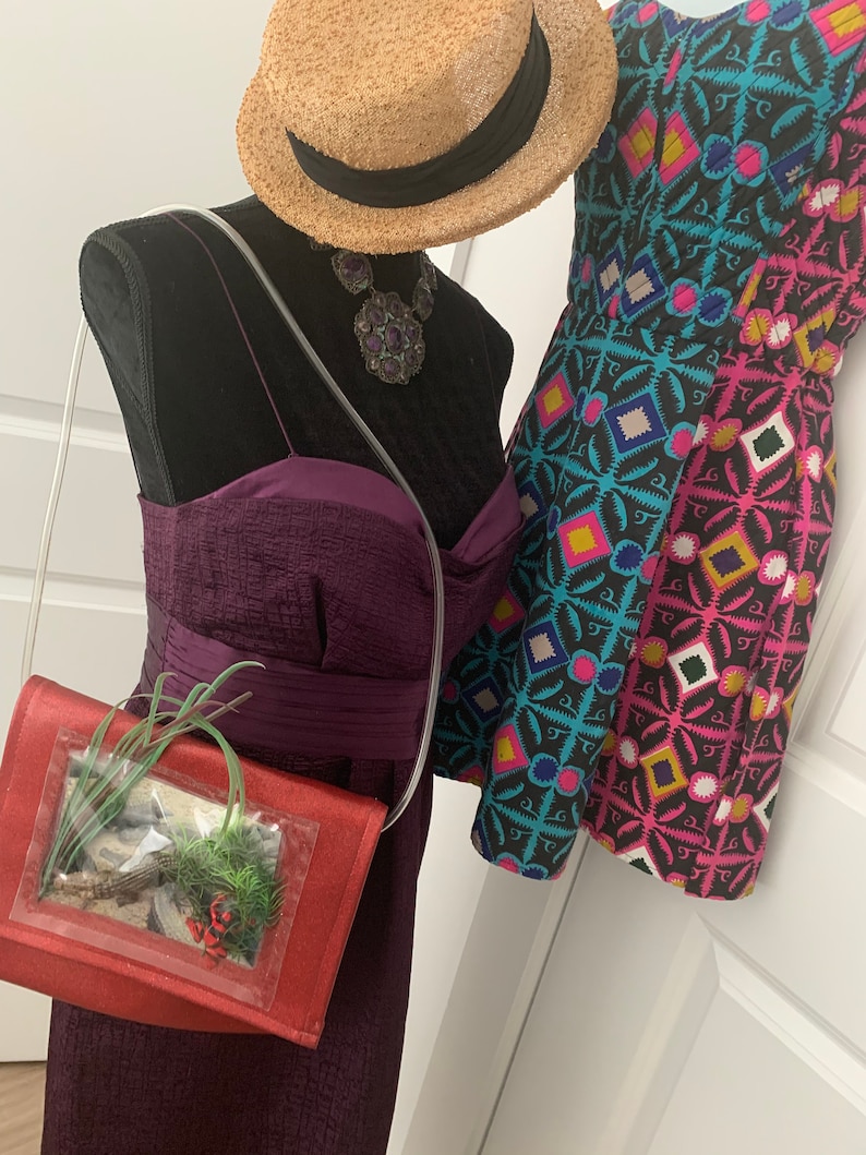 May include: A red purse with a clear plastic window showing a small potted plant inside. The purse is on a mannequin wearing a purple dress and a straw hat with a black band. The mannequin is also wearing a large, dark purple and silver necklace.
