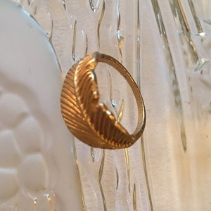 May include: A gold-toned ring with a feather design. The feather is textured and has a detailed design.