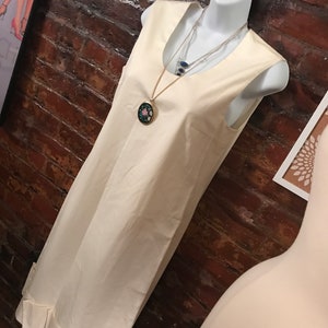 May include: A sleeveless, ivory-colored dress with a ruffled hem. The dress is on a white mannequin and has a gold chain necklace with a floral pendant.