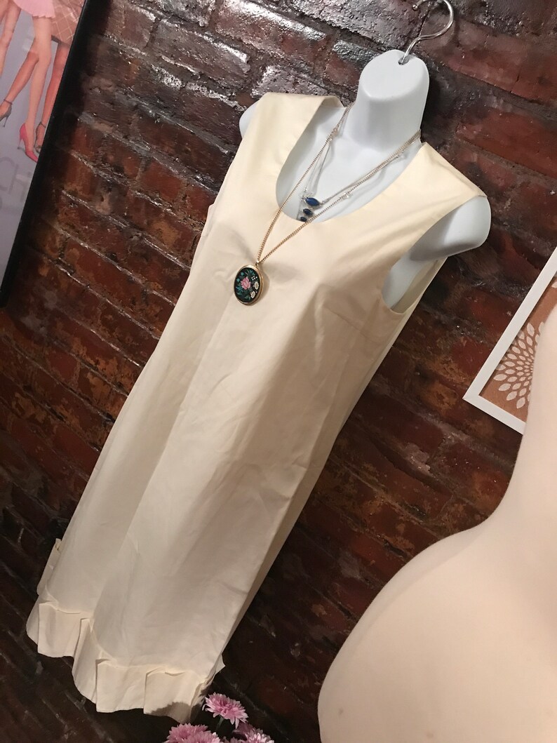 May include: A sleeveless, ivory-colored dress with a ruffled hem. The dress is on a white mannequin and has a gold chain necklace with a floral pendant.