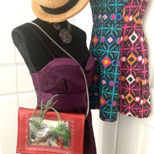 May include: A red purse with a clear plastic window, showcasing a small potted plant inside. The purse has a long strap and is worn on a mannequin dressed in a purple dress and a straw hat.