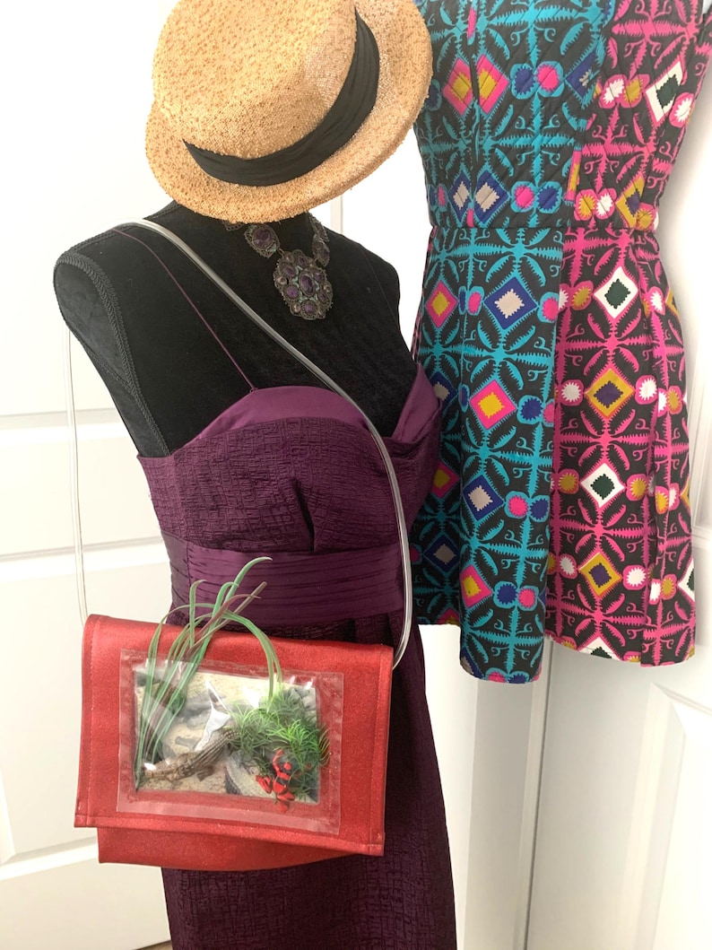 May include: A red purse with a clear plastic window, showcasing a small potted plant inside. The purse has a long strap and is worn on a mannequin dressed in a purple dress and a straw hat.