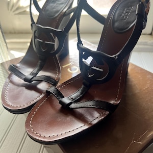 May include: A pair of black leather sandals with a T-strap design featuring a circular Gucci logo cutout. The sandals have a brown leather footbed with white contrast stitching, an adjustable ankle strap with a metal buckle, and a block heel.