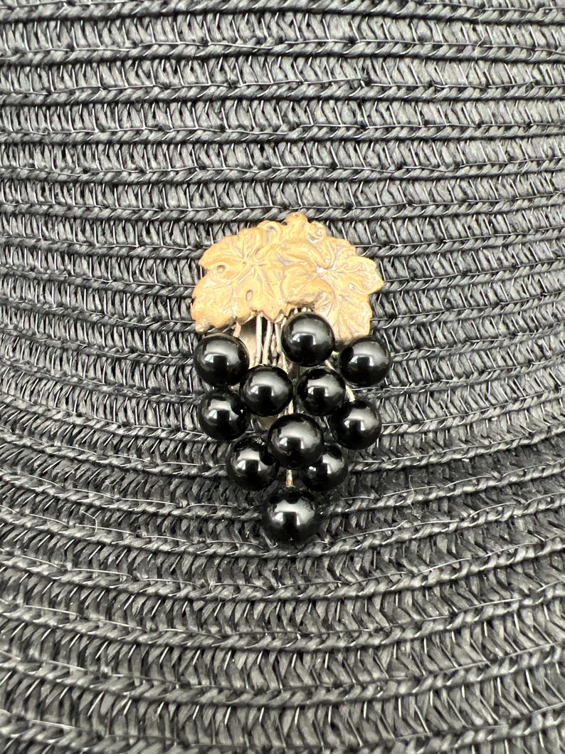 May include: A black and gold grape cluster brooch with a detailed floral design. The brooch is made of metal and has a cluster of black beads that resemble grapes.