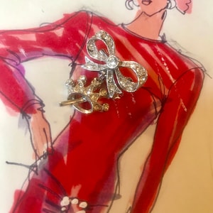 May include: A silver and crystal bow brooch with a crown charm. The brooch is pinned to a red and white striped dress on a fashion illustration.