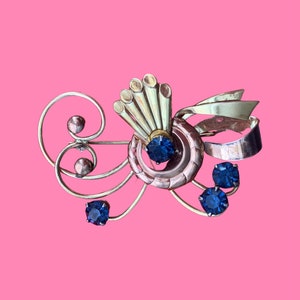 May include: A gold and silver tone brooch with a fan-shaped design and blue rhinestones. The brooch has a delicate, intricate design.