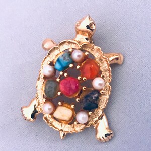 May include: A gold-toned turtle brooch with a shell made of colorful stones and pearls. The turtle's shell features a variety of colors, including blue, red, and green. The brooch has a textured, detailed design.