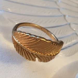 May include: A gold-toned ring with a feather design. The feather is detailed with lines and has a textured surface.