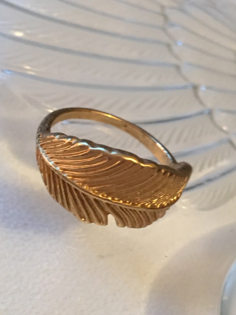 May include: A gold-toned ring with a feather design. The feather is detailed with lines and has a textured surface.