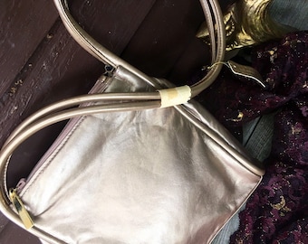 May include: A metallic gold-toned leather tote bag with a zipper closure and two top handles. The bag is sitting on a dark purple and gold fabric.