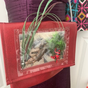 May include: A red glitter clutch bag with a clear plastic window featuring a small alligator and a red and black frog. The bag has a green plant design on the front.