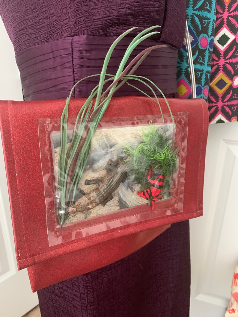 May include: A red glitter clutch bag with a clear plastic window featuring a small alligator and a red and black frog. The bag has a green plant design on the front.
