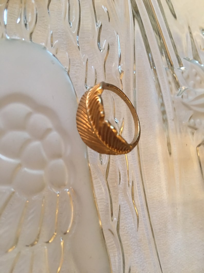 May include: A gold ring with a feather design. The ring is on a white background.