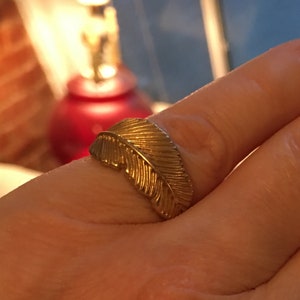 May include: A gold ring with a feather design. The ring is on a person's finger.